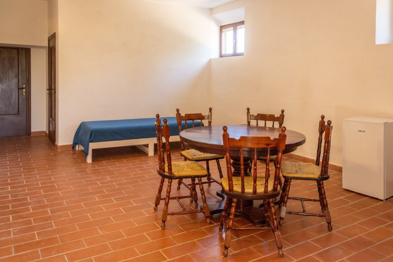 A dining area with a round table and six chairs, a small refrigerator, a bed with a blue cover, and a window.