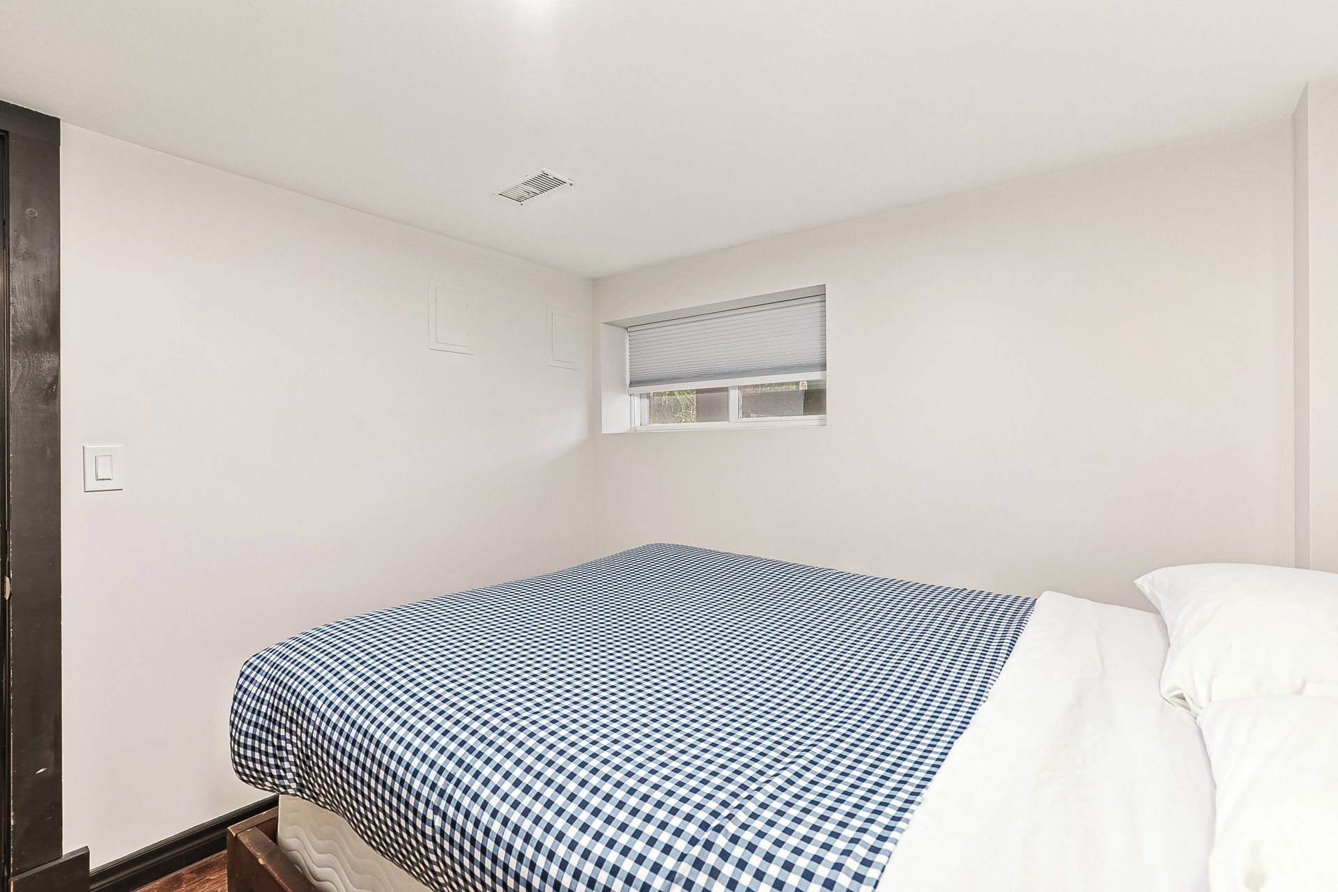 A small, single-bed room with a checkered bedspread, a window with blinds, and a white wall.