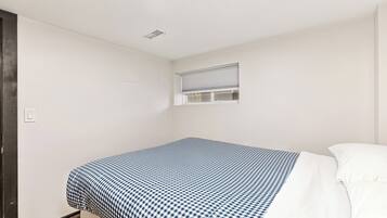 A small, single-bed room with a checkered bedspread, a window with blinds, and a white wall.
