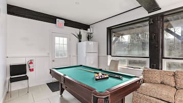 A pool table with balls, a sofa, a door with an exit sign, a refrigerator, and a window with a view of trees.