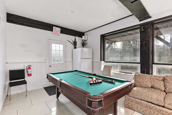 Une table de billard avec des billes, un canapé, une porte avec un panneau indiquant la sortie, un réfrigérateur et une fenêtre donnant sur des arbres.