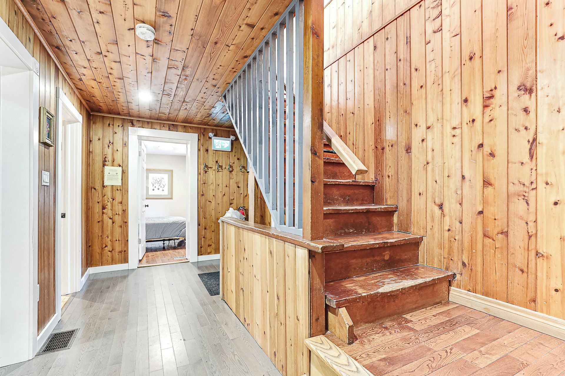 A wooden interior with a staircase, a door leading to a room with a checkered bed, and a small hallway.