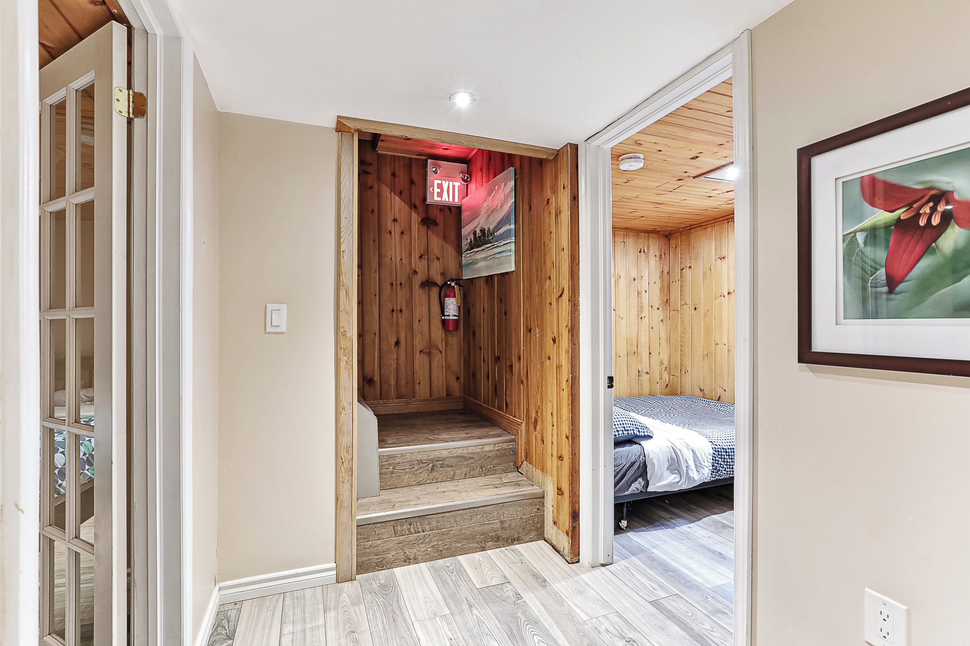 A hallway leading to a bedroom with a bed, wooden walls, a fire extinguisher, and a door with an exit sign.