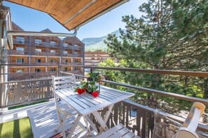 Ein Balkon mit Tisch und Stühlen, der auf ein Gebäude mit zahlreichen Fenstern und eine Berglandschaft blickt.