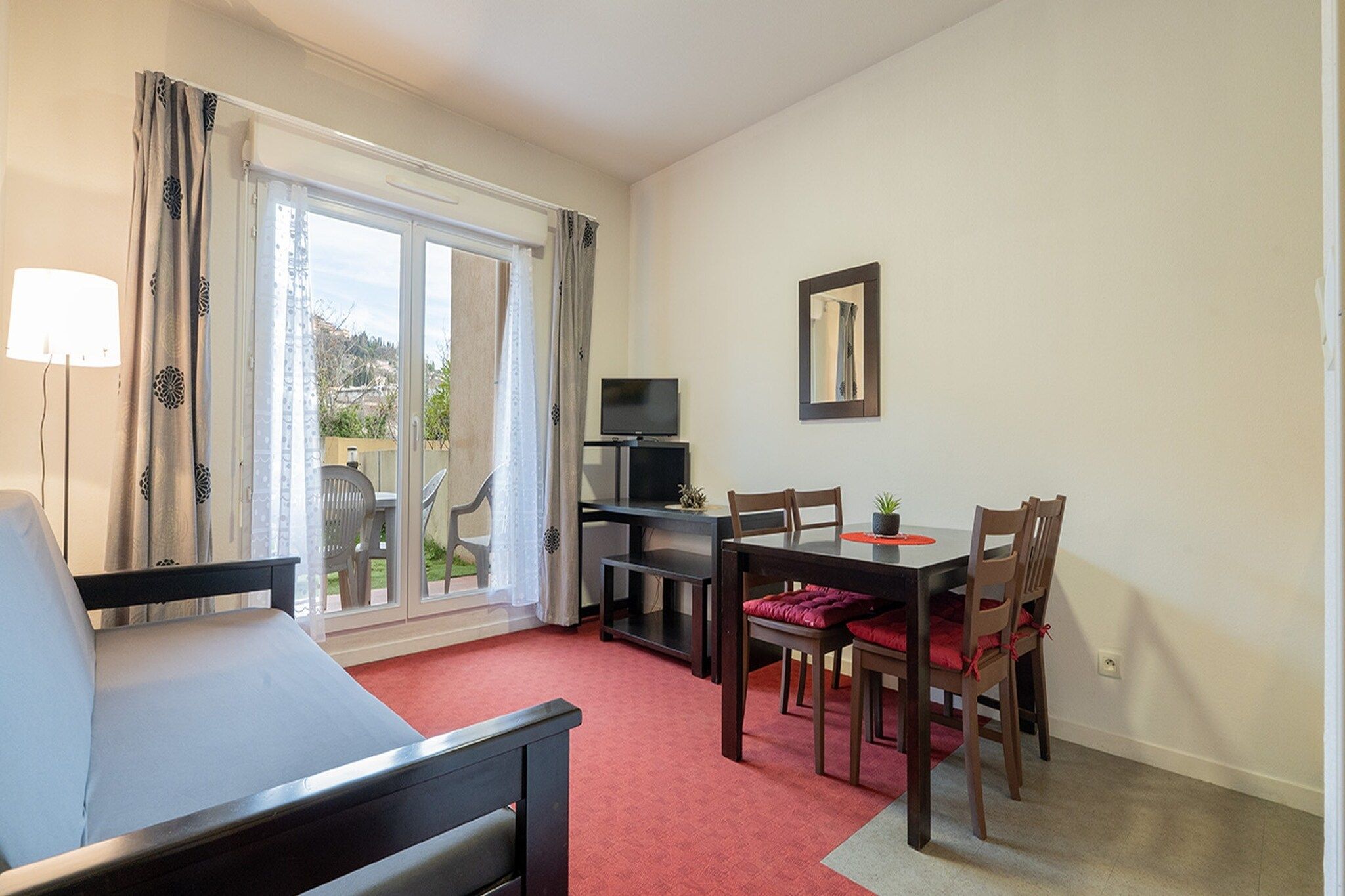 A room with a bed, a dining table with chairs, a TV, and a sliding glass door leading to a balcony.