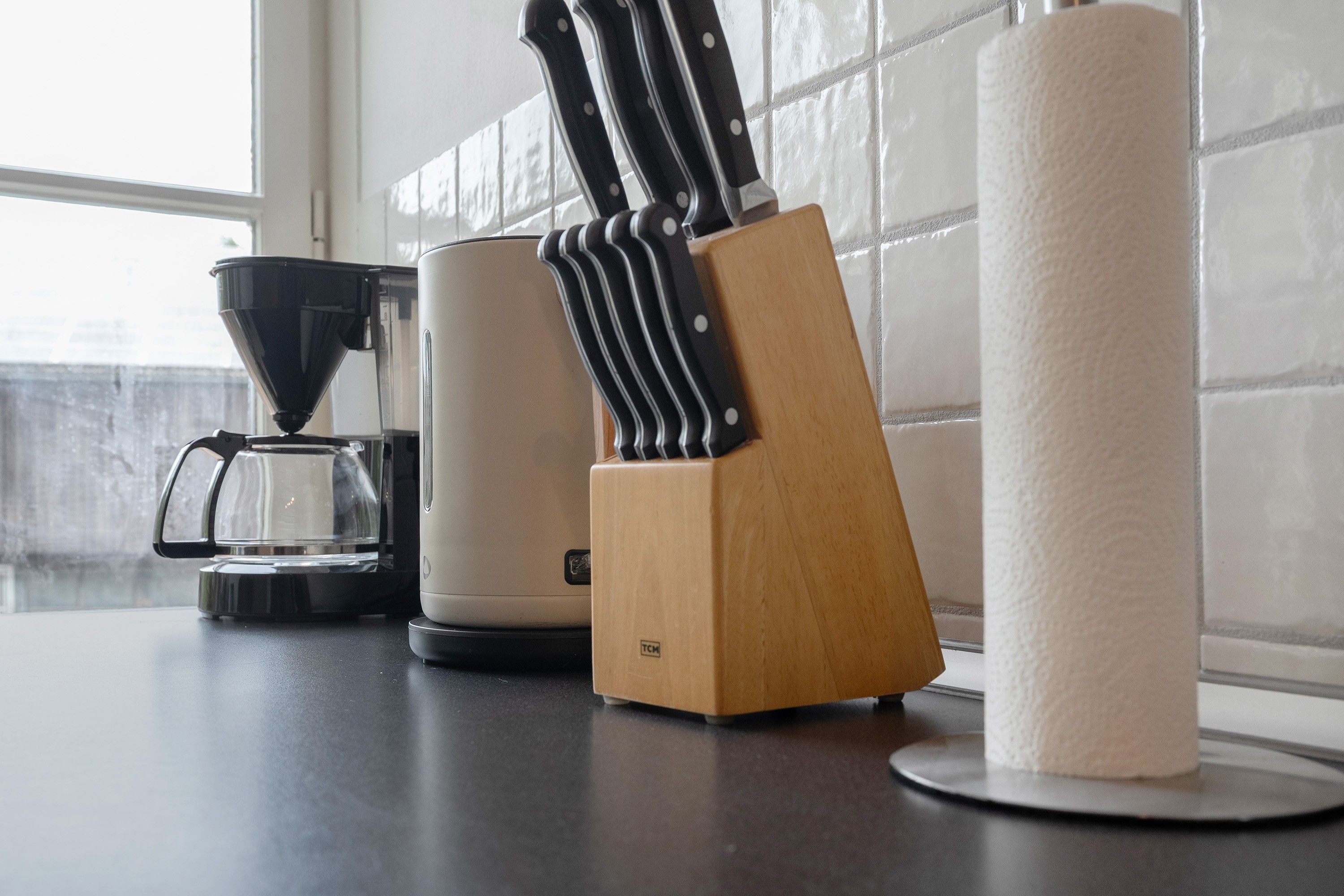 Un taulell de cuina amb una cafetera, un suport per ganivets i un rotlle de paper de cuina.