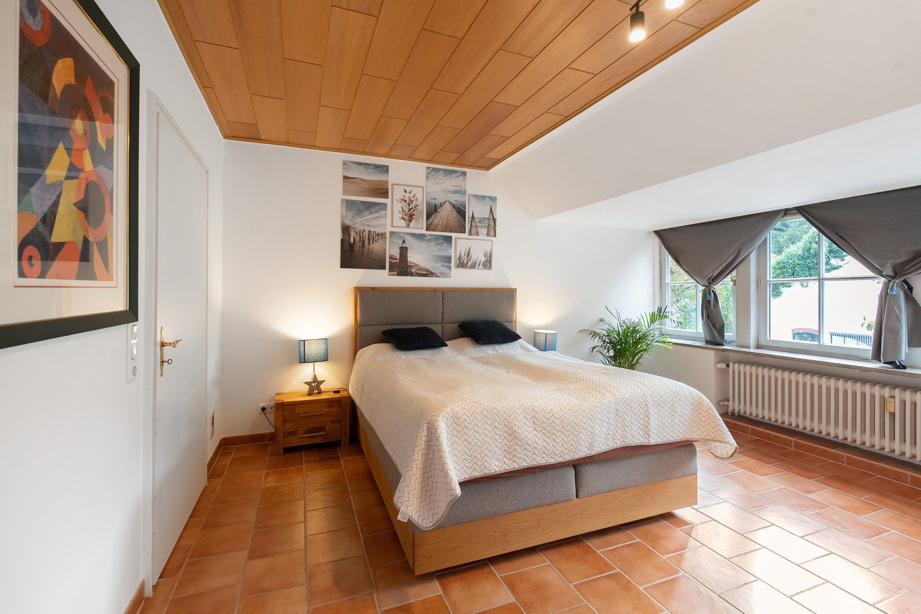 Ein Schlafzimmer mit Holzboden, einem Bett mit grauem Kopfteil, einem Nachttisch mit Lampe und einem Fenster mit Vorhängen.