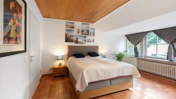 Un dormitorio con techo de madera, una cama con cabecera gris, una mesita de noche con lámpara y una ventana con cortinas.