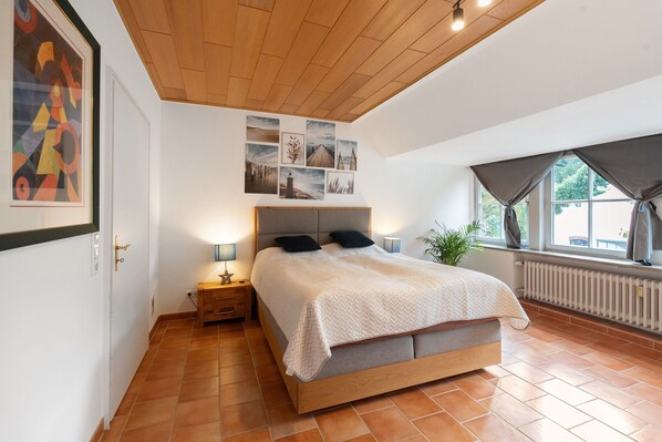 A bedroom with a wooden ceiling, a bed with a grey headboard, a nightstand with a lamp, and a window with curtains.
