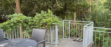 An outdoor patio with a round table and wicker chairs, surrounded by a metal railing and lush greenery.
