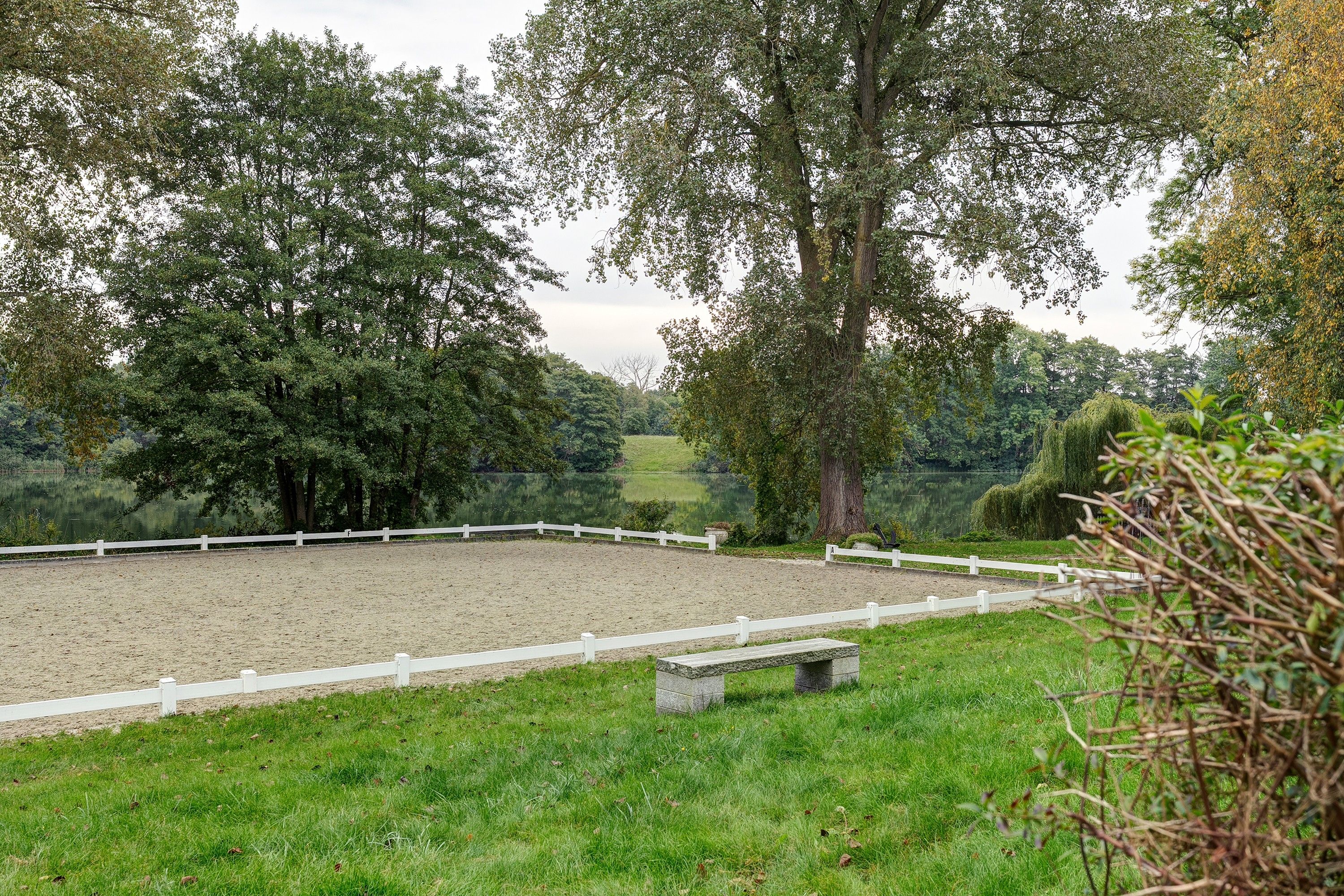 A fenced equestrian arena with a bench, surrounded by trees and a body of water.