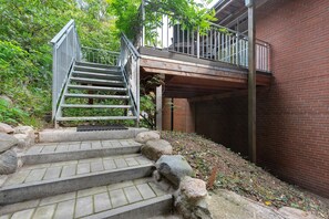 A set of stone steps leading to a metal staircase with a wooden deck above, surrounded by greenery and a brick wall.