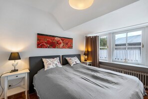 A bedroom with a bed, two bedside lamps, a nightstand, a wall-mounted painting, and a window with curtains.