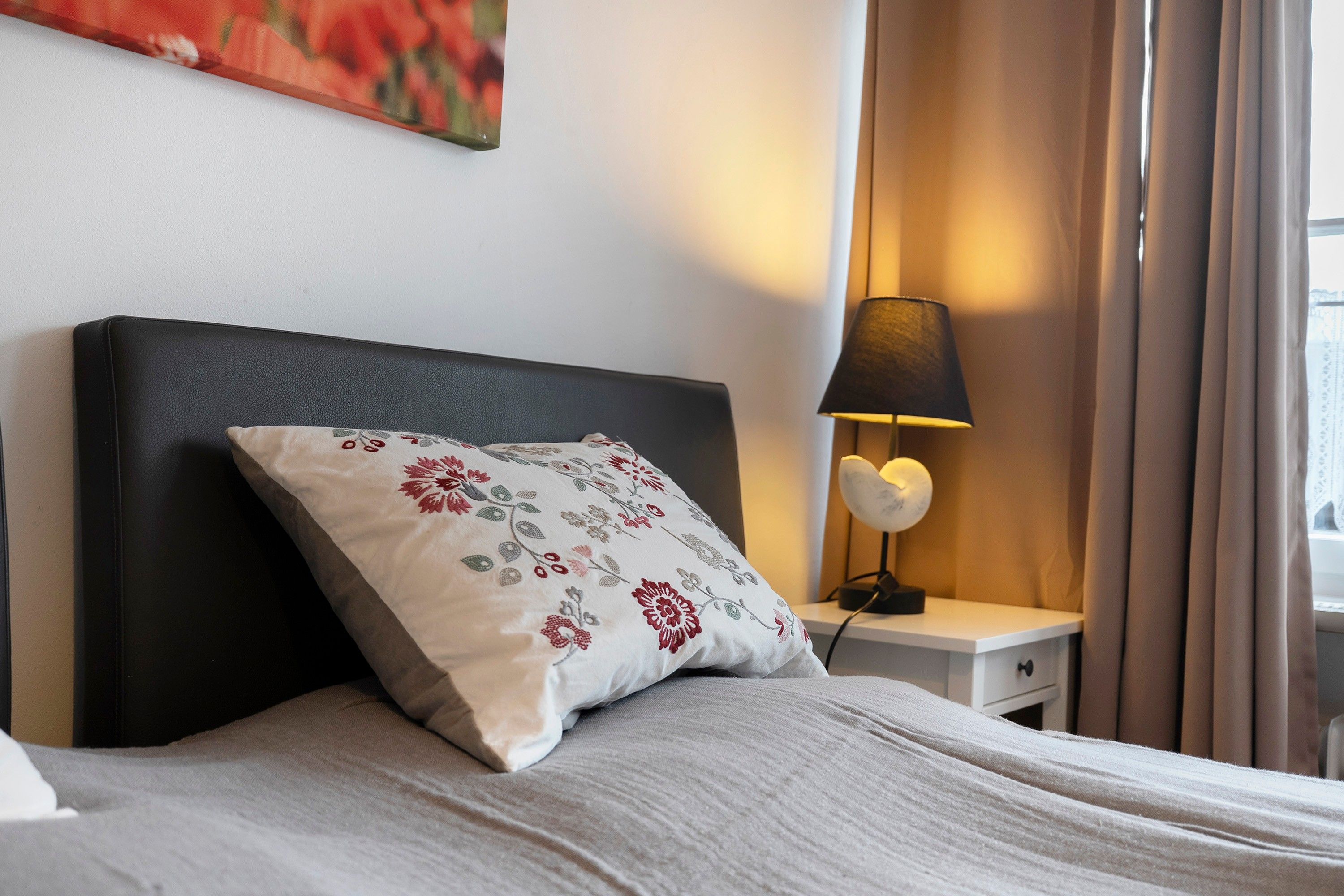 Een keurig opgemaakt bed met een bloemenkussen, een bedlampje en een schilderij aan de muur.