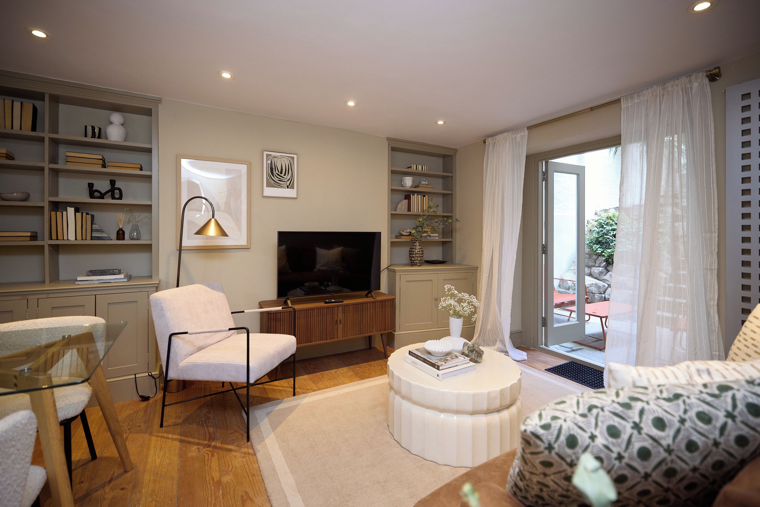 A modern living room with a flat-screen TV, a white armchair, a sofa, a coffee table, and a bookshelf.
