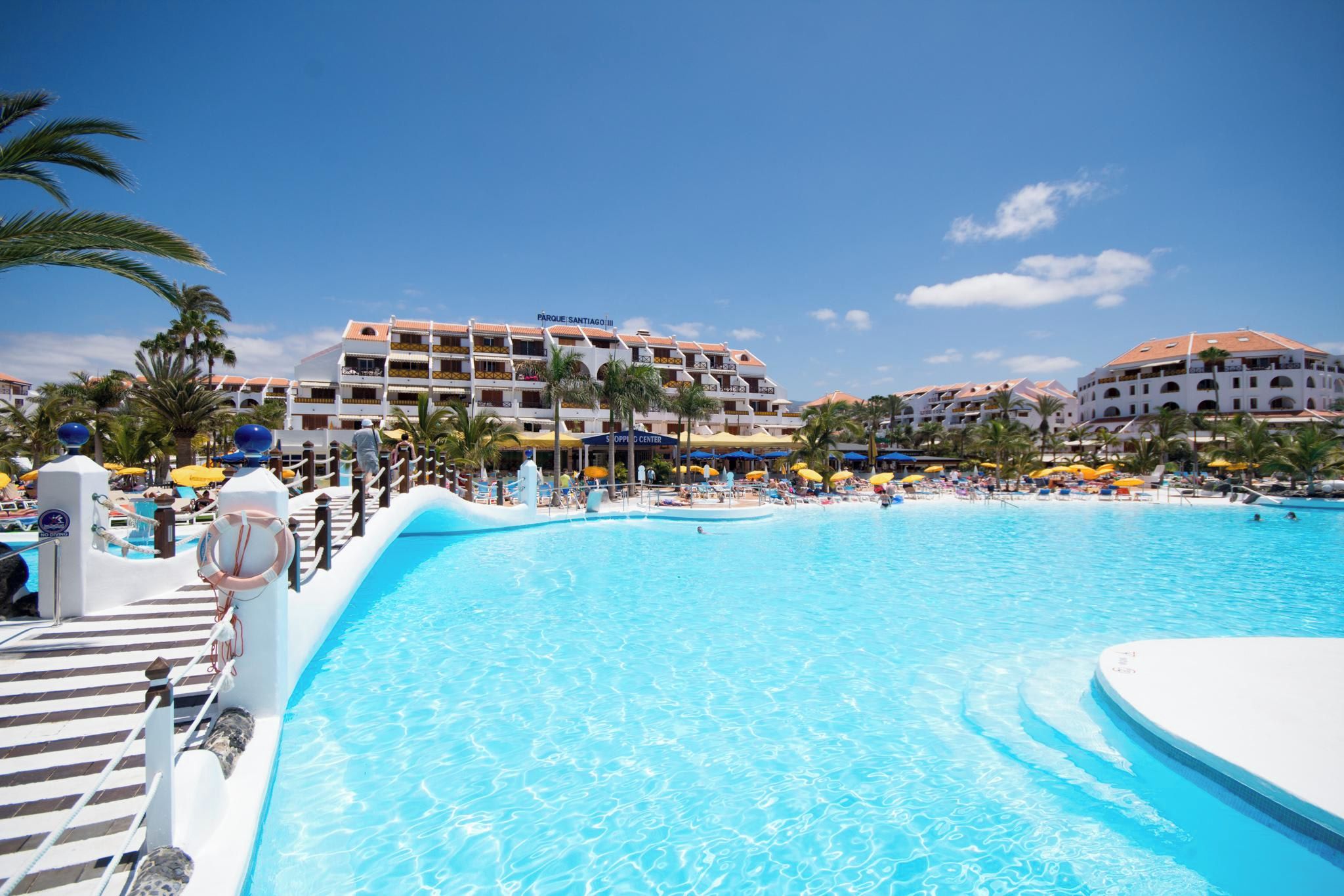 Una gran piscina con agua cristalina y azul, rodeada de palmeras y sombrillas.