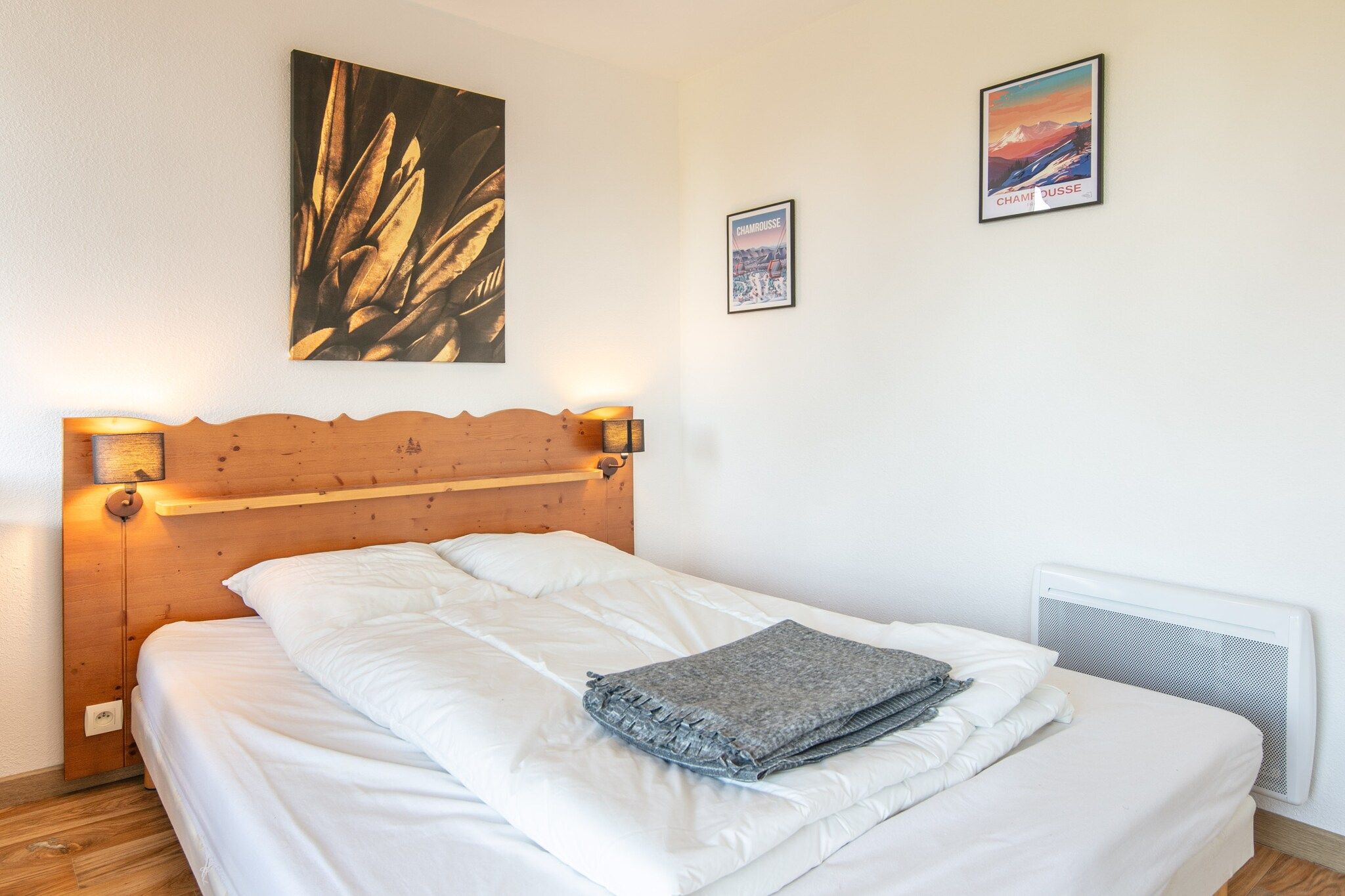 A neatly made bed with white linens and a folded blanket, a wooden headboard with wall-mounted lights, and two framed pictures on the wall.