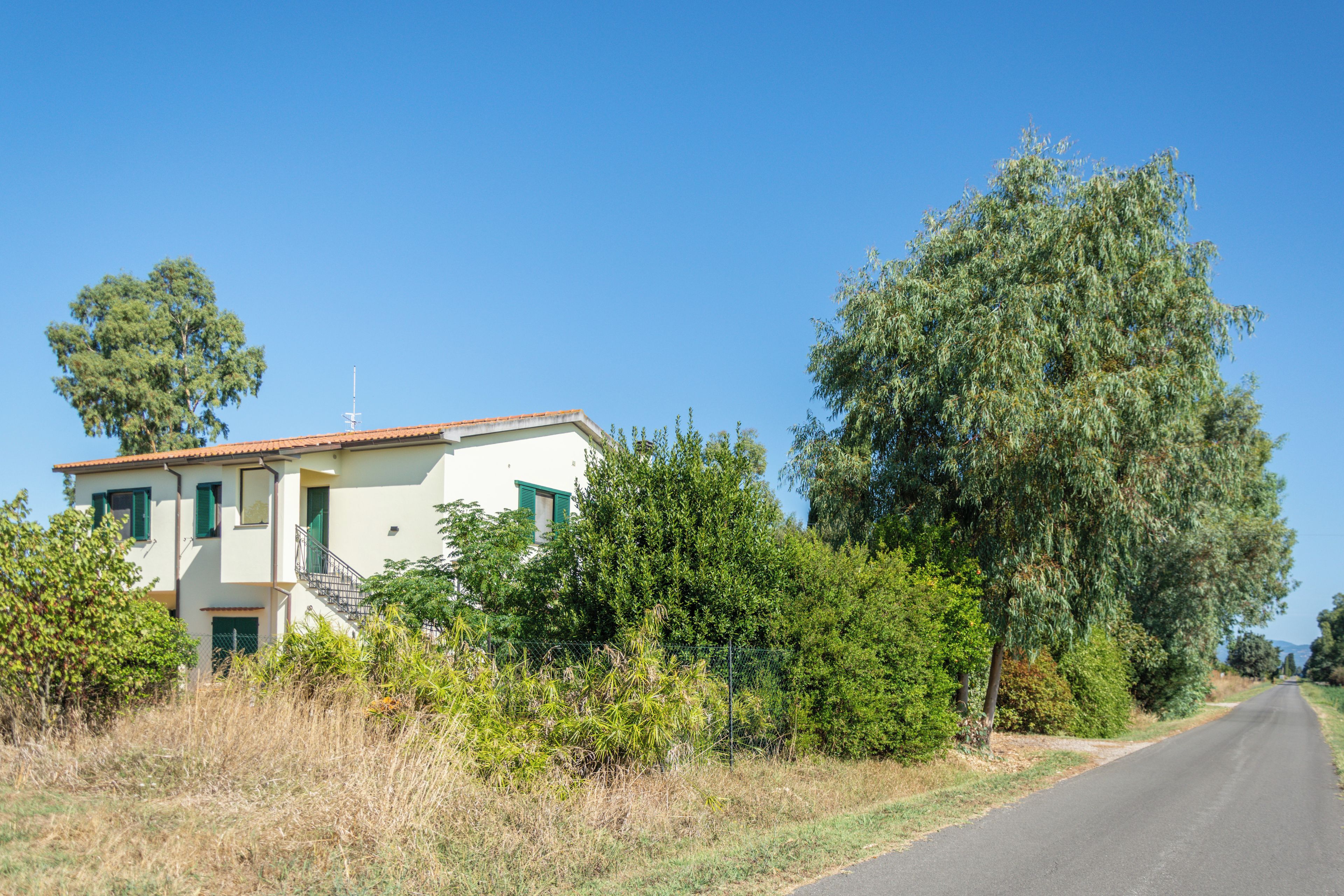 A two-story building with a white facade and green shutters, surrounded by dense greenery and a clear sky.