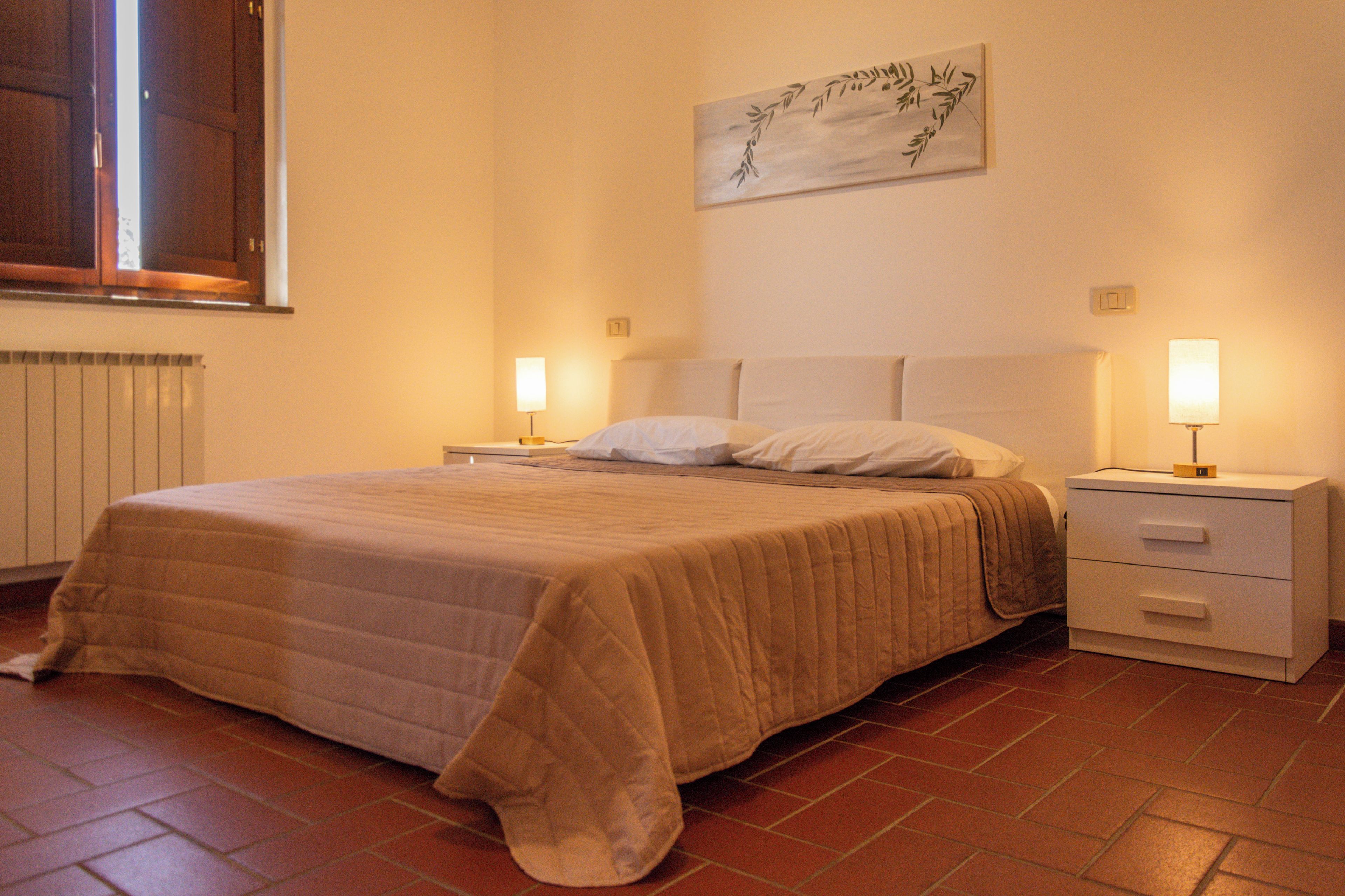 A neatly made bed with a beige blanket, a bedside table with a lamp, and a wall-mounted artwork.