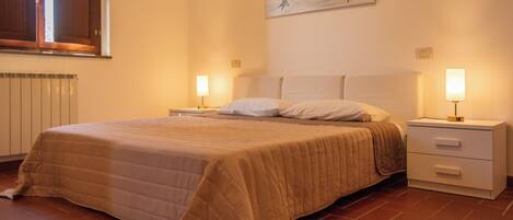 A neatly made bed with a beige blanket, a bedside table with a lamp, and a wall-mounted artwork.