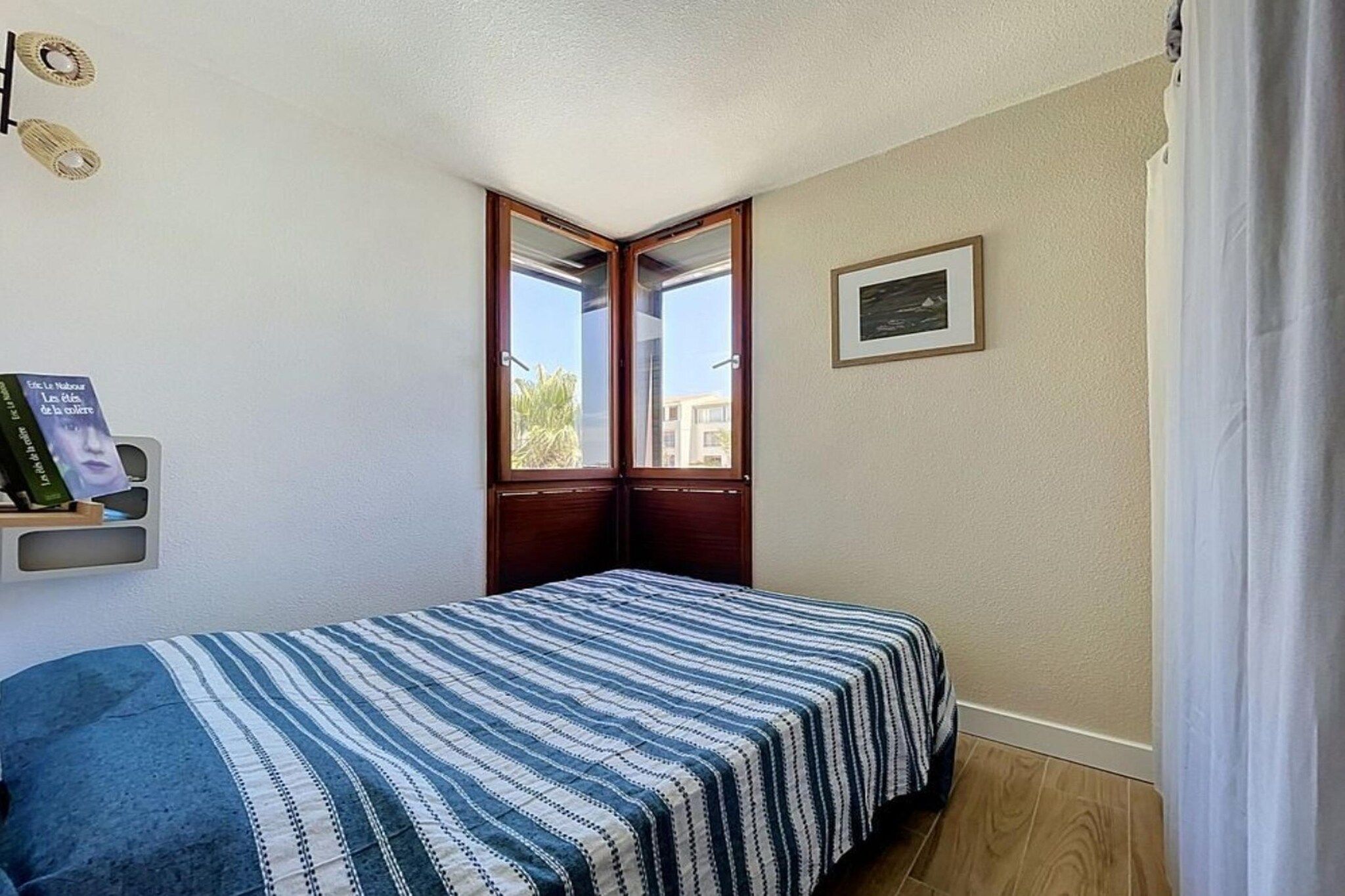 A bedroom with a bed, a window, a framed picture, and a shelf with books.