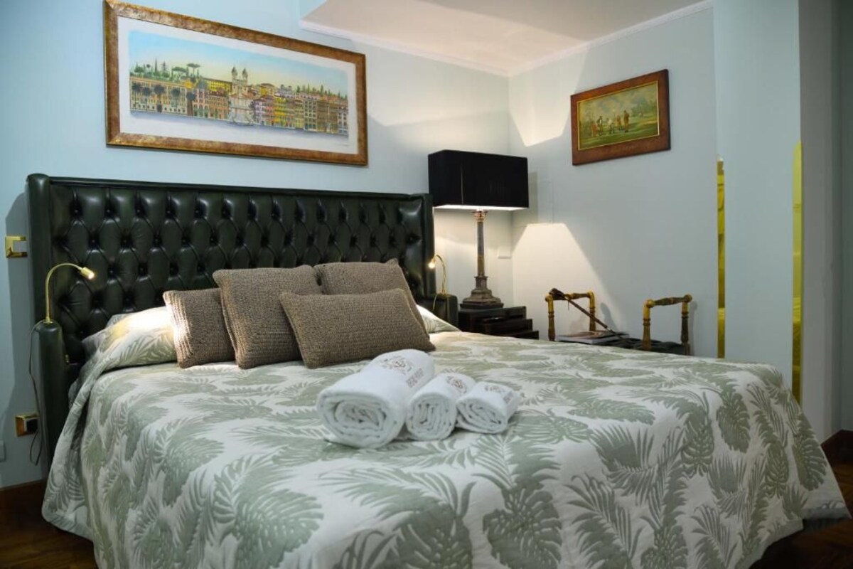 A hotel room with a large bed, a headboard, a painting on the wall, a lamp, and a small table with a lamp.