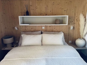 Ein Schlafzimmer mit Holzwand, einem weißen Bett mit zwei Kissen, einem Nachttisch mit Lampe und einem Regal mit Dekoration.