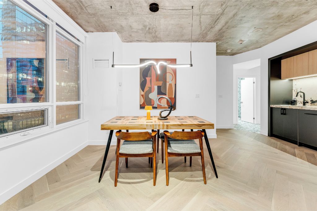 A modern kitchen with a wooden table and chairs, a view through large windows, and a concrete ceiling.