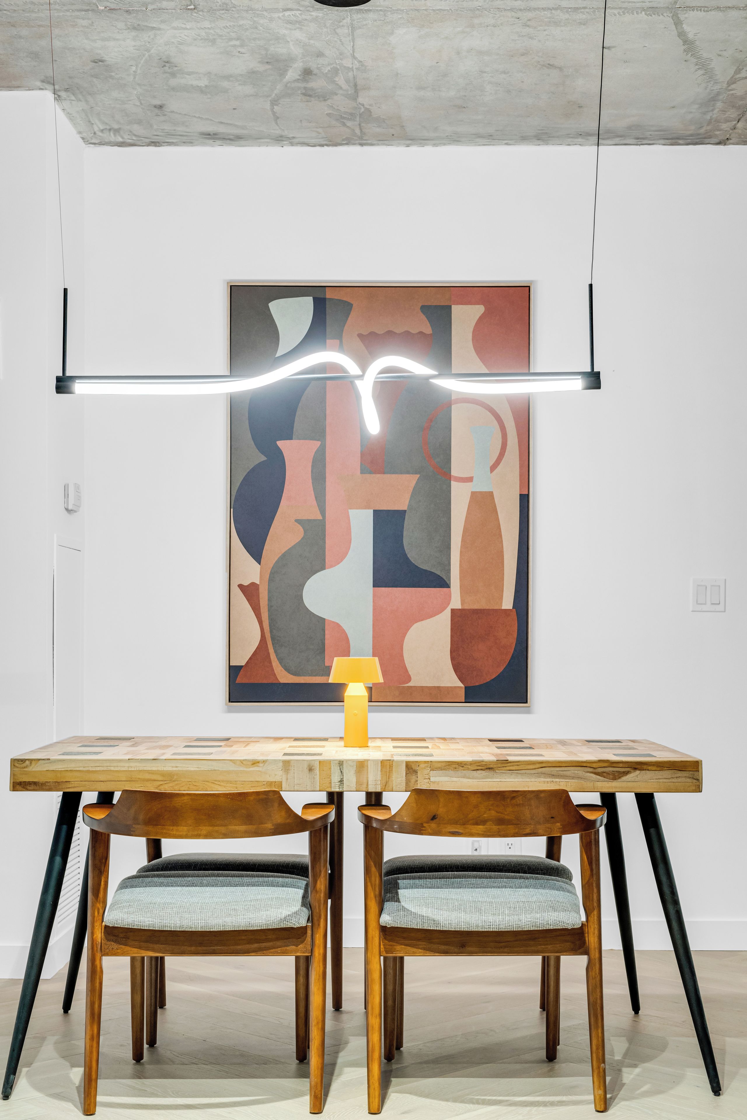 A modern dining area with a wooden table and chairs, a contemporary abstract painting on the wall, and a pendant light above.