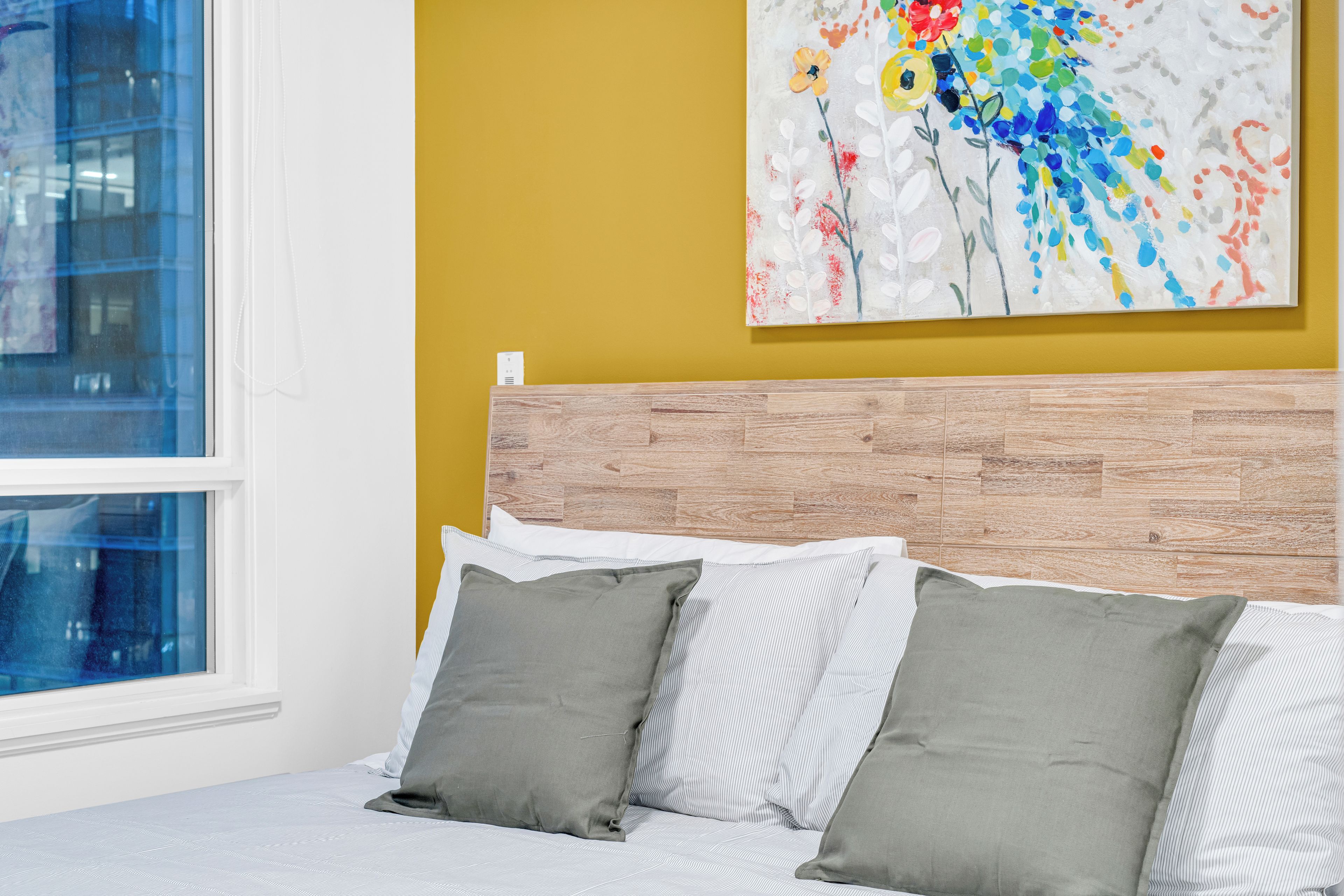 A bedroom with a bed, pillows, a wooden headboard, a window, and a colorful abstract painting on the wall.