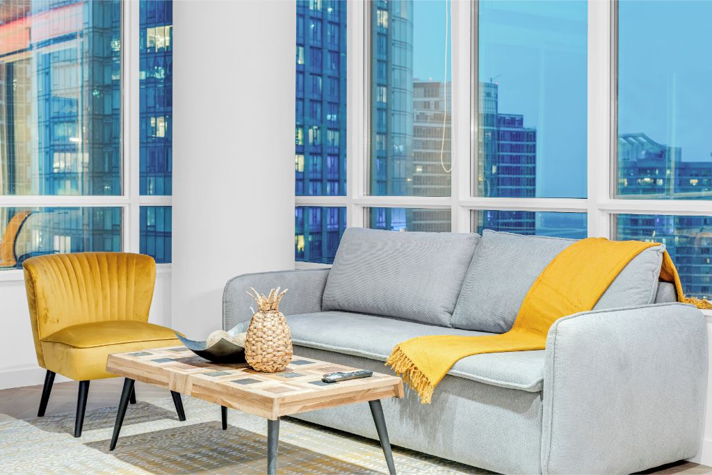 A modern living room with a grey sofa, a yellow armchair, and a wooden coffee table with a pineapple and remote control.