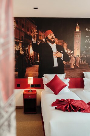 A hotel room with a large poster of a man in traditional attire, a bed with white and red bedding, a small bedside table with a lamp, and a red nightstand. - Art Hotel (Marrakech)