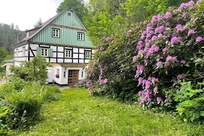 A traditional half-timbered house with a green roof and white walls, surrounded by lush greenery and flowering bushes. - Charming Apartment with Mountain Views in Nordenau for 6 (Nordenau)