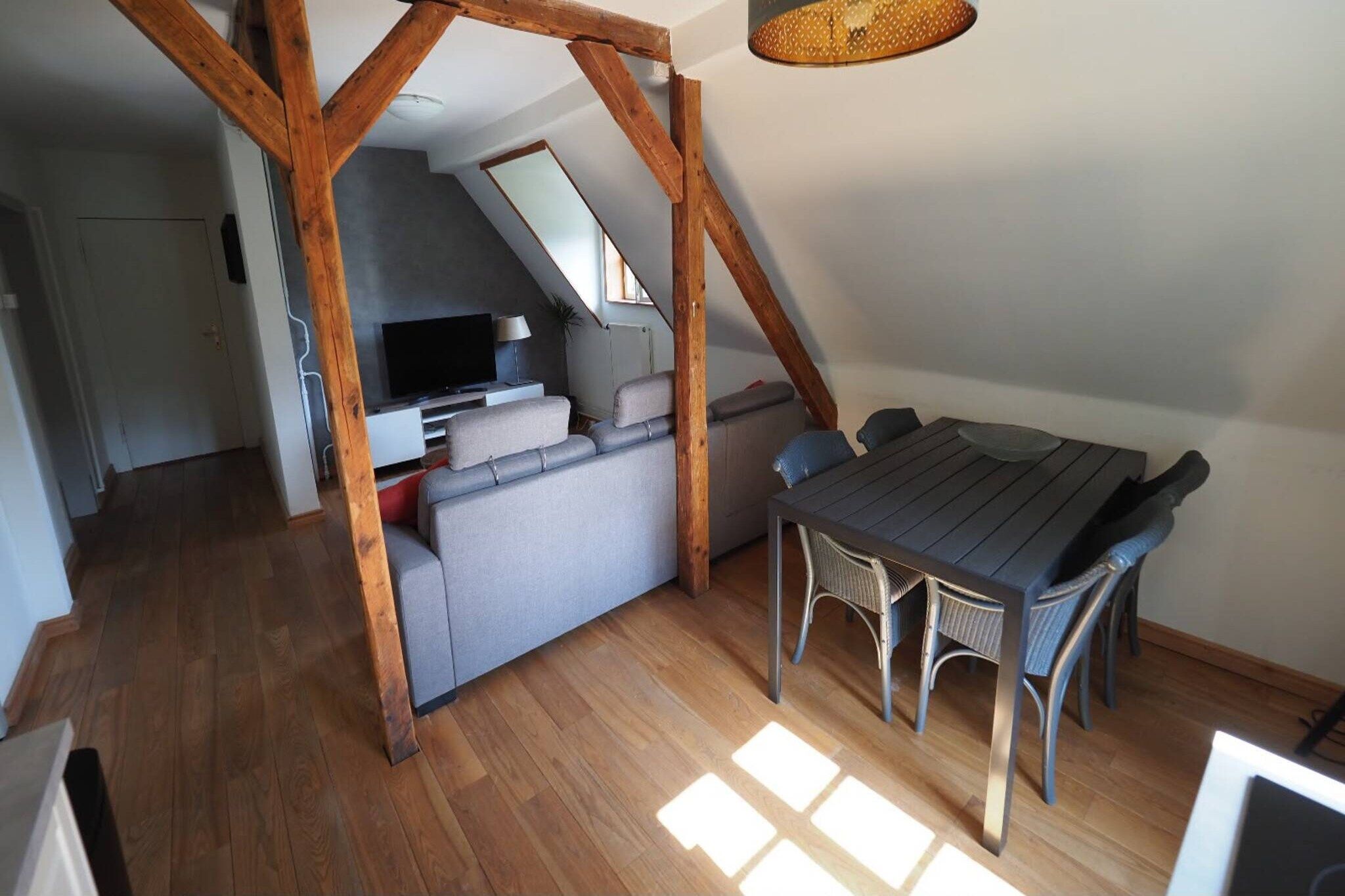 Un cuarto estilo loft con techo de vigas de madera, una mesa de comedor con sillas, un sofá gris y un televisor al fondo.