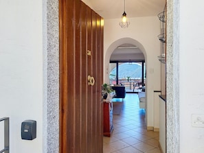 Un pasillo con puerta de madera, suelo de baldosas, un mostrador de recepción y vista a un balcón con una montaña al fondo.