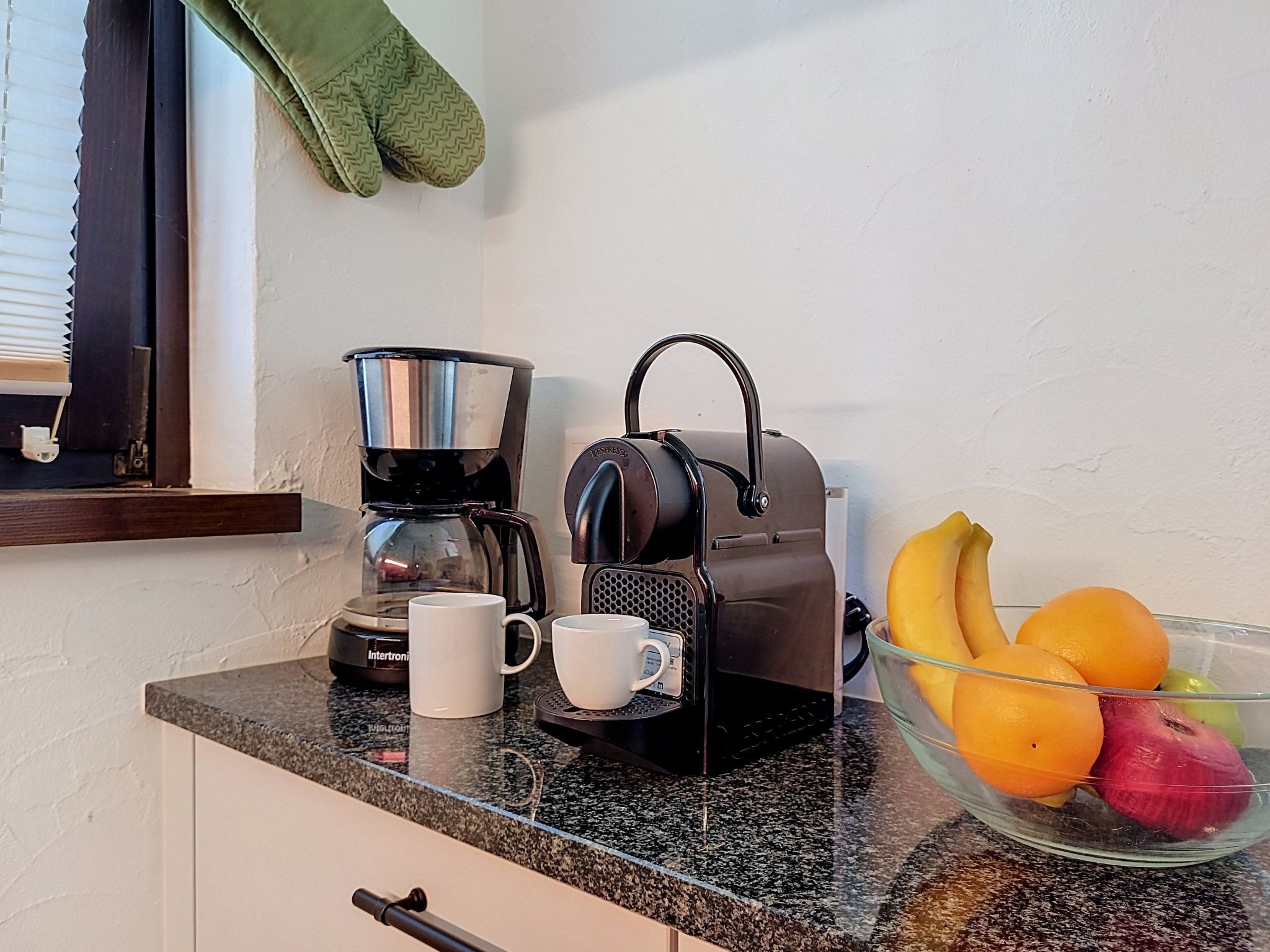 En köksbänk med en kaffebryggare, en mugg, en fruktskål med bananer och äpplen och ett fönster med persienner.