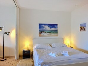 A hotel room with a bed, two bedside lamps, a nightstand, a small rug, and a painting of a beach scene. - Residenza Miralago Apt.D31 by Interhome (Piazzogna)