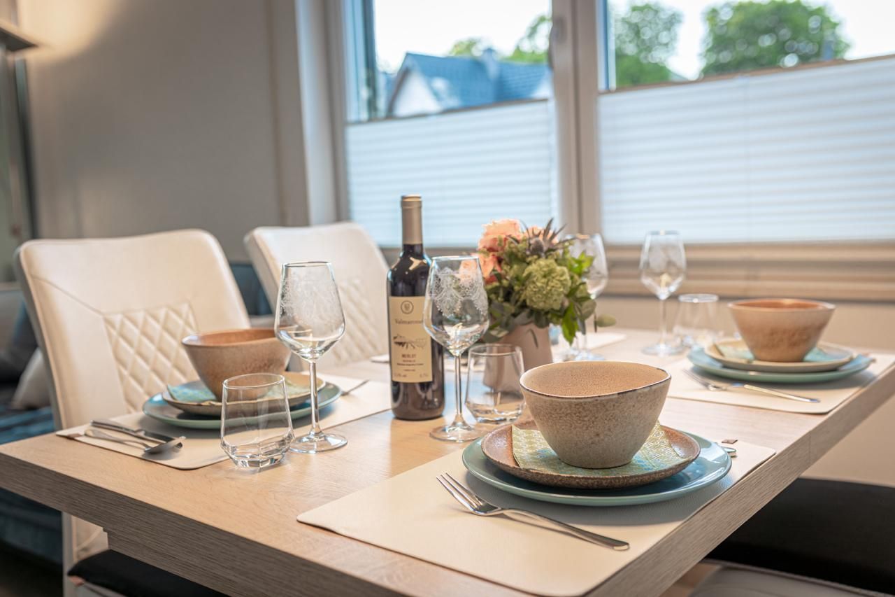 A dining table set with bowls, wine glasses, and a bottle, with a window view in the background.