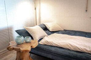 A bed with a blue mattress and white bedding, a small wooden table with two decorative vases, and a window with blinds. - SOLIM: Kitchen, balcony & garden, 45 square meters, max. 4 people. (Kiefersfelden)