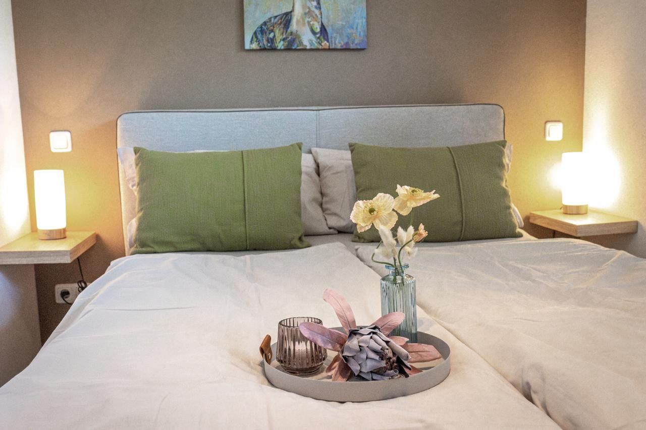 Ein ordentlich bezogenes Bett mit einem Tablett voller Blumen und Kerzen, zwei grüne Kissen und ein Gemälde an der Wand.