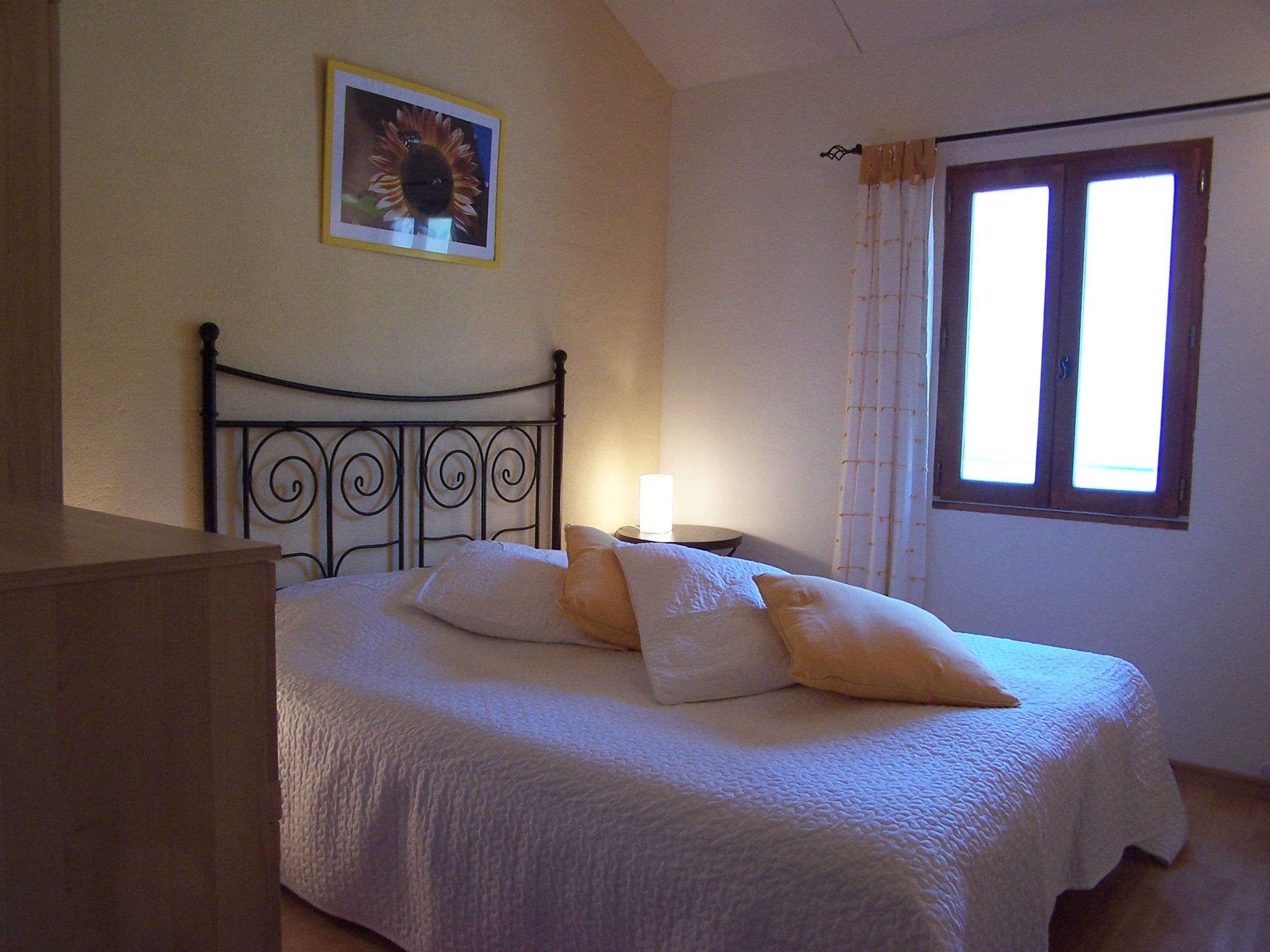 A bedroom with a bed, pillows, a nightstand, a lamp, a window with curtains, and a framed picture on the wall.