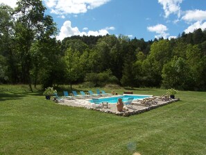 Ein Schwimmbecken mit Liegestühlen und einer Steingrenze, umgeben von einer Rasenfläche und Bäumen.