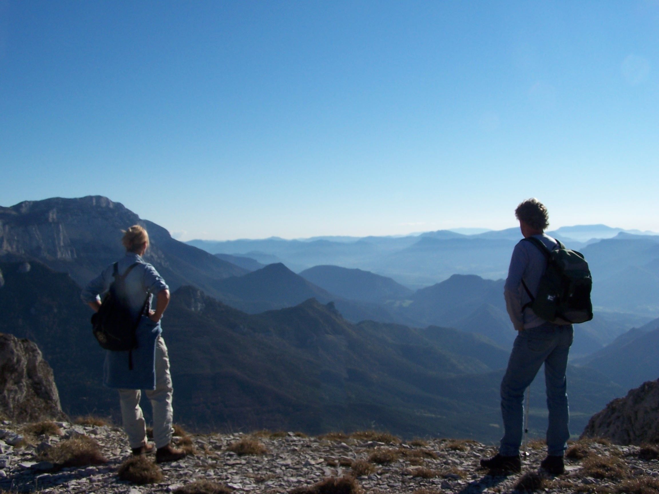 Dos personas de pie en la cima de una montaña rocosa, con vista a montañas distantes.