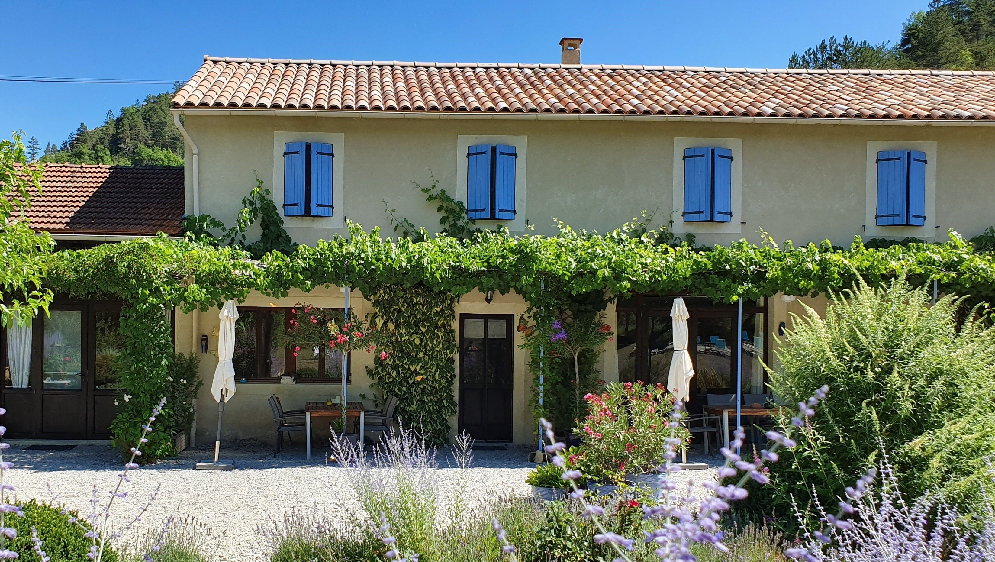 A two-story building with a tiled roof, blue shutters, and a covered garden area with seating.