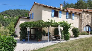 A two-story house with a stone facade, blue shutters, and a covered patio area with outdoor furniture. - Gîte "Cigale" with Pool, Minimum Age 18 Years (Beaumont-en-Diois)