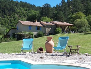 A pool area with two blue chairs, a large terracotta pot, and a stone building in the background. - Gîte "Cigale" with Pool, Minimum Age 18 Years (Beaumont-en-Diois)