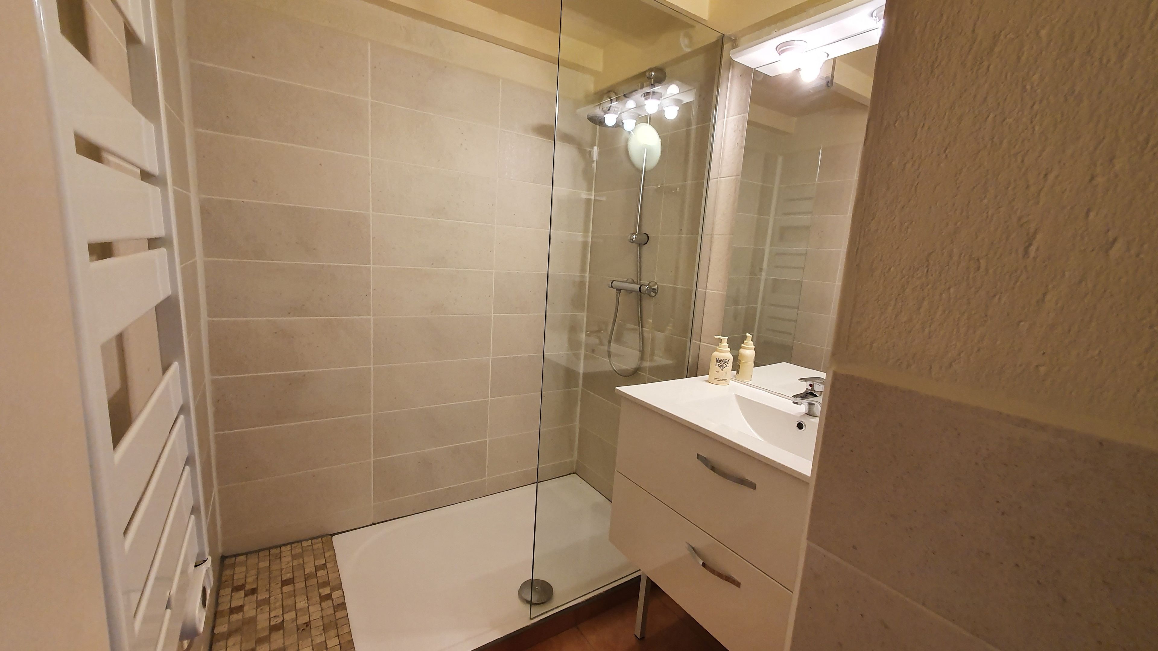 A bathroom with a glass shower enclosure, a white sink with a mirror, and a tiled floor.