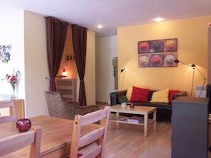 Ein Wohnzimmer mit einer Couch, einem Esstisch und einem Sessel. Auf dem Tisch steht eine Lampe und auf der Couch ein Blumentopf.