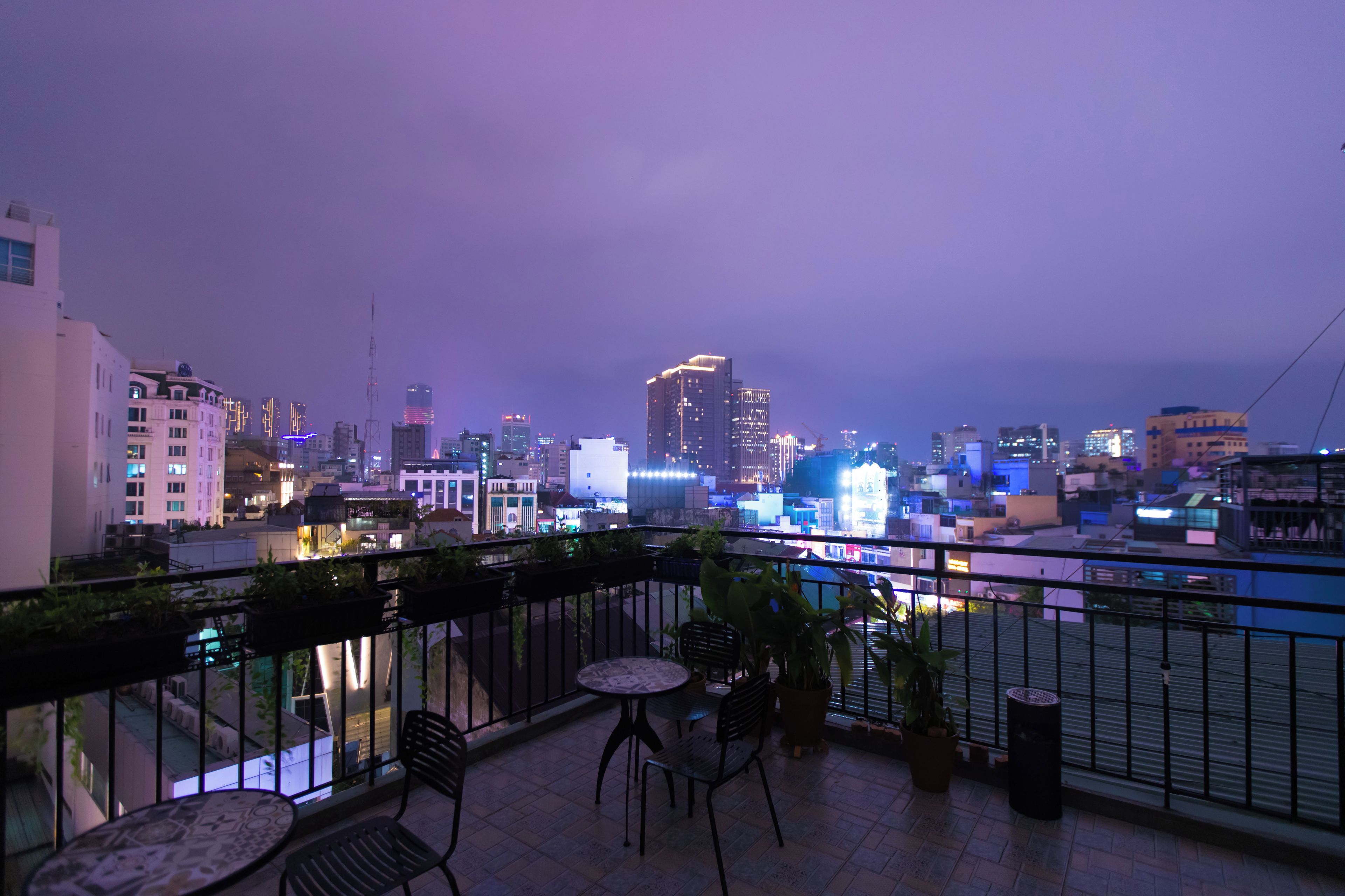 A balcony with chairs and a table, overlooking a cityscape with illuminated buildings at dusk.