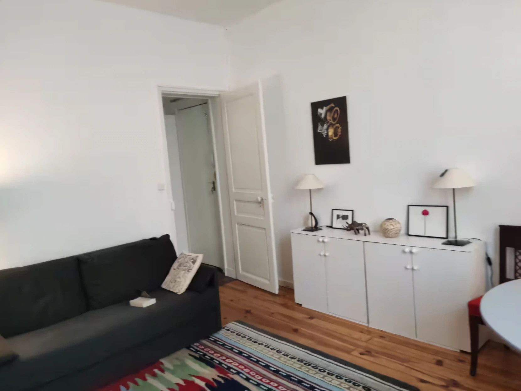 Een woonkamer met een donkergrijze bank, een witte kast met decoratieve objecten, een kamerkleed met dessin, en een deur naar een andere kamer.